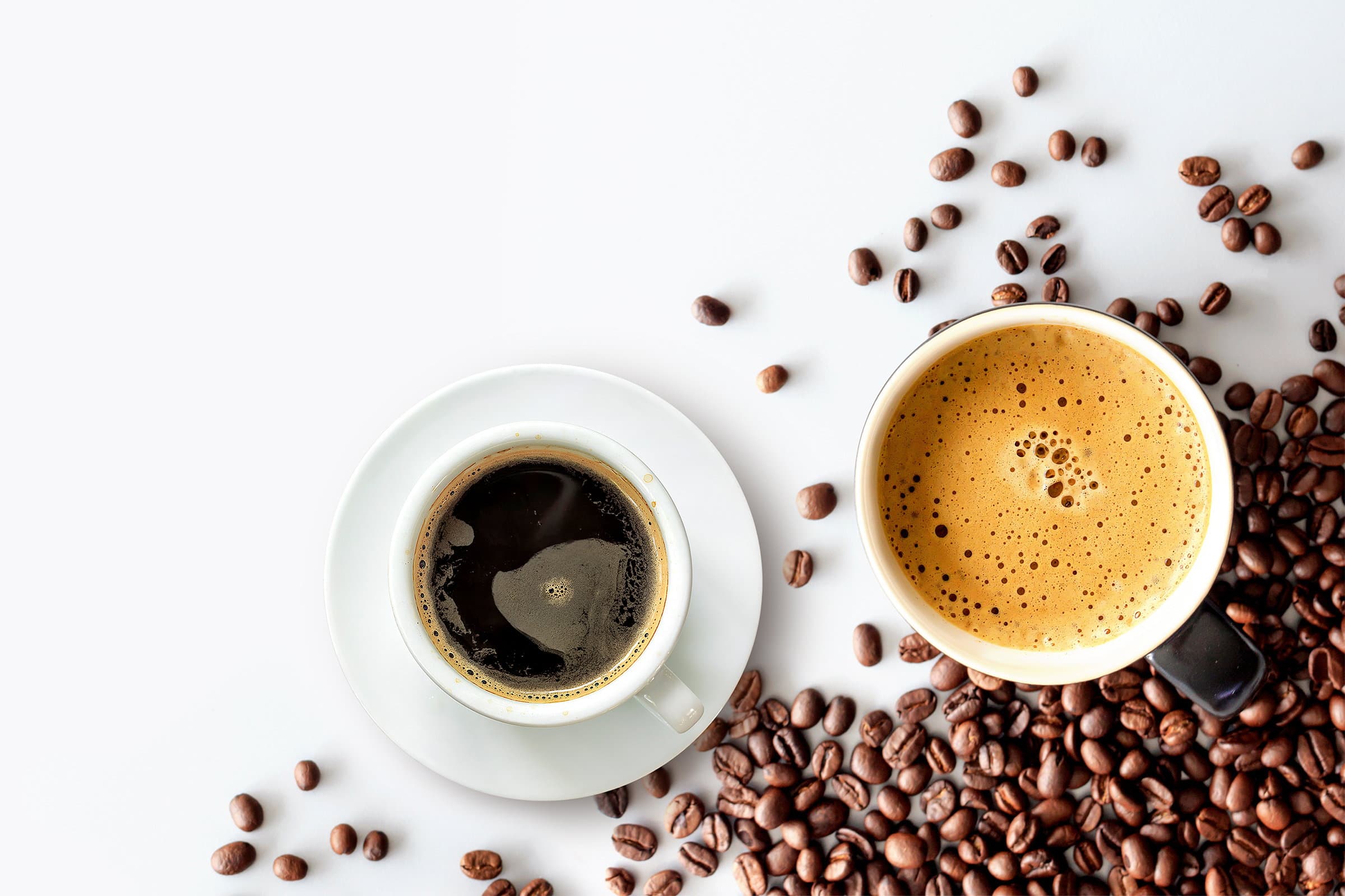 A cup of black filter coffee and a cup of espresso with a thick crema, photographed from above on white with roasted beans scattered around them.
