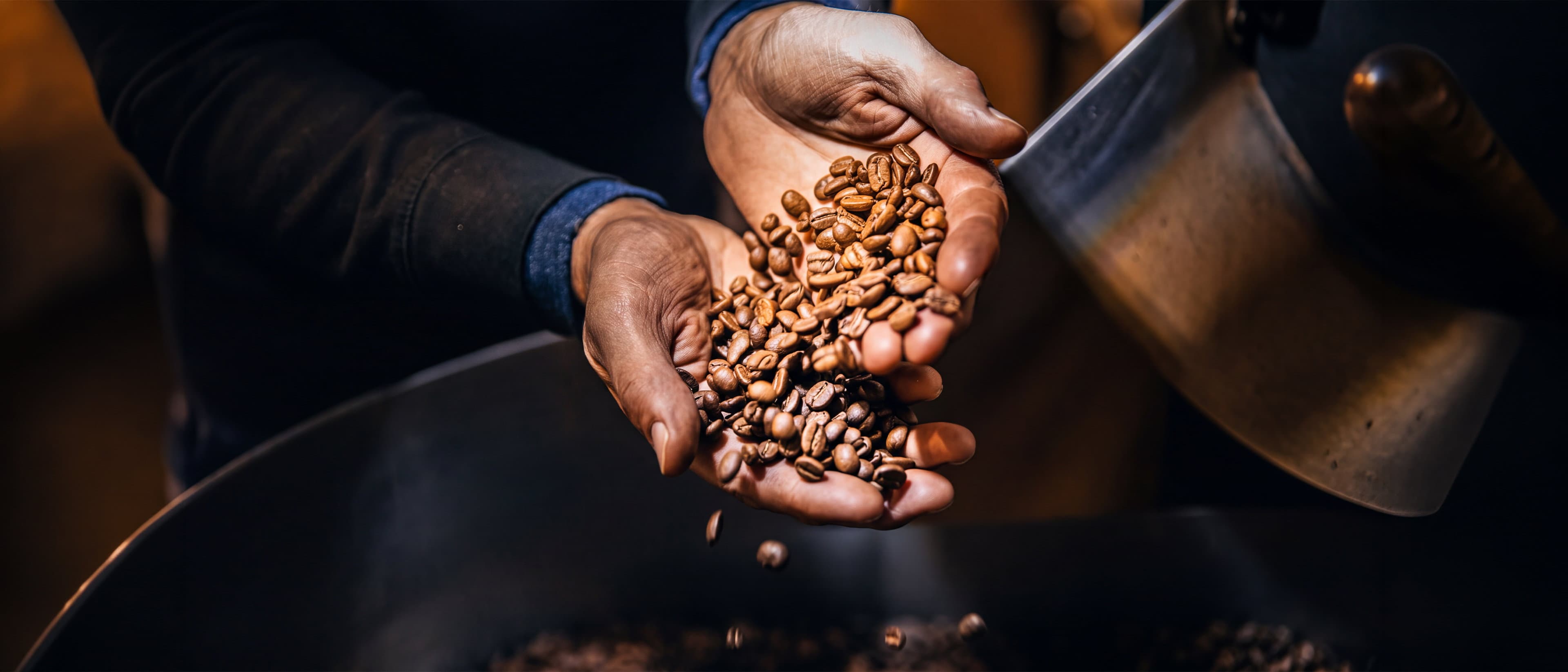 Two hands cupping freshly roasted coffee beans beside the open drum of a roaster.