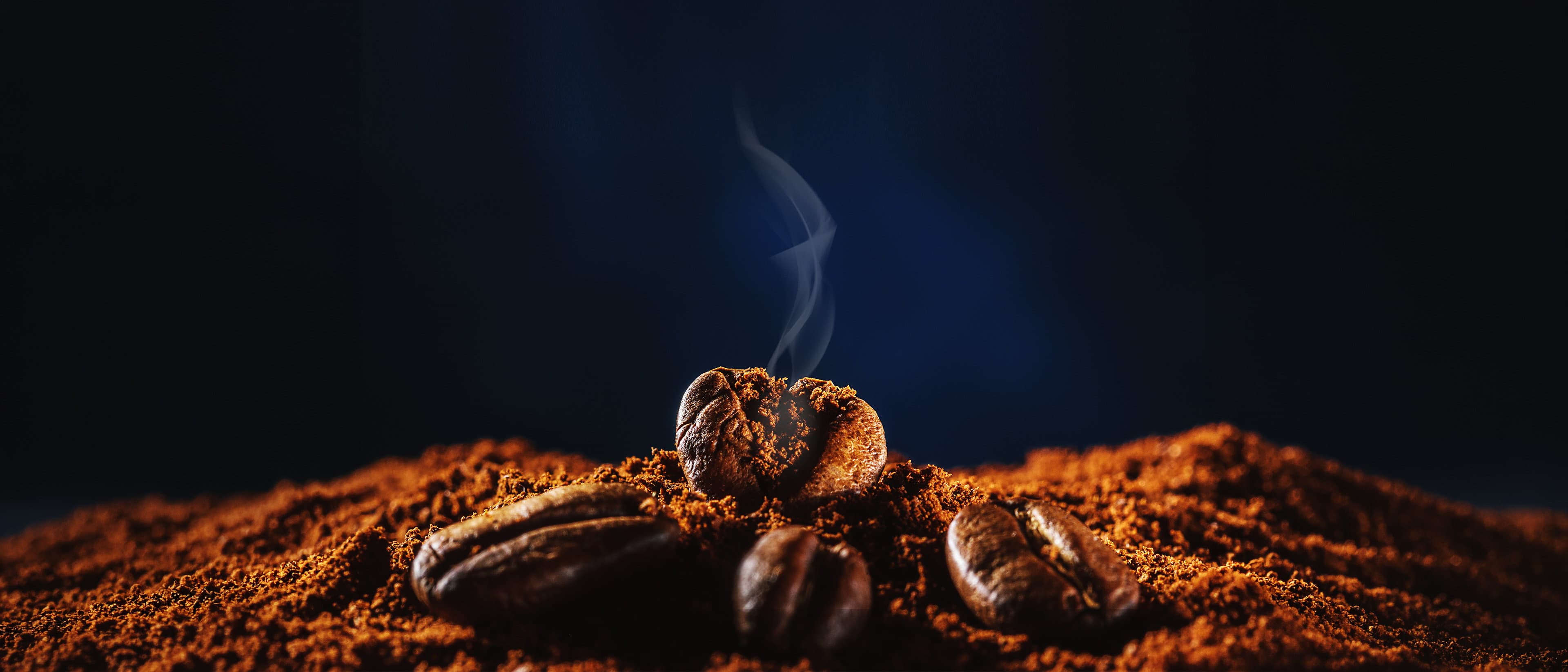 A mound of freshly ground coffee with whole roasted beans resting on it, lit against a dark background with steam rising.